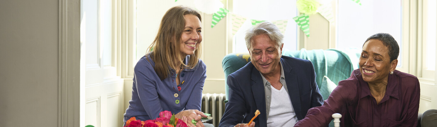 Three people are sitting down in a lounge. In front of them is a table decorated for Coffee Morning, full of fruit, sweet treats and cups of warm drinks. They appear to be talking and eating food together.