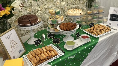 A table is decorated for a Coffee Morning. It has different sweet and savoury treats on it that are labelled. Behind the table is a white rose flower wall. On the wall is a poster about testicular cancer.