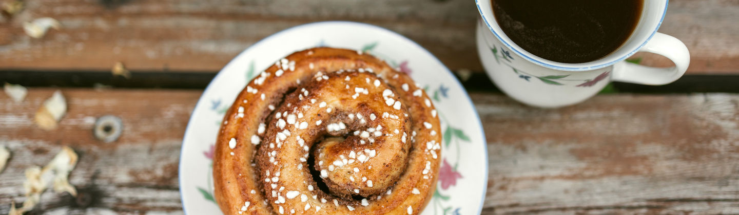 A small plate with a traditional cinnamon bun. Next to it is a black mug of coffee. They are both on a wooden table.