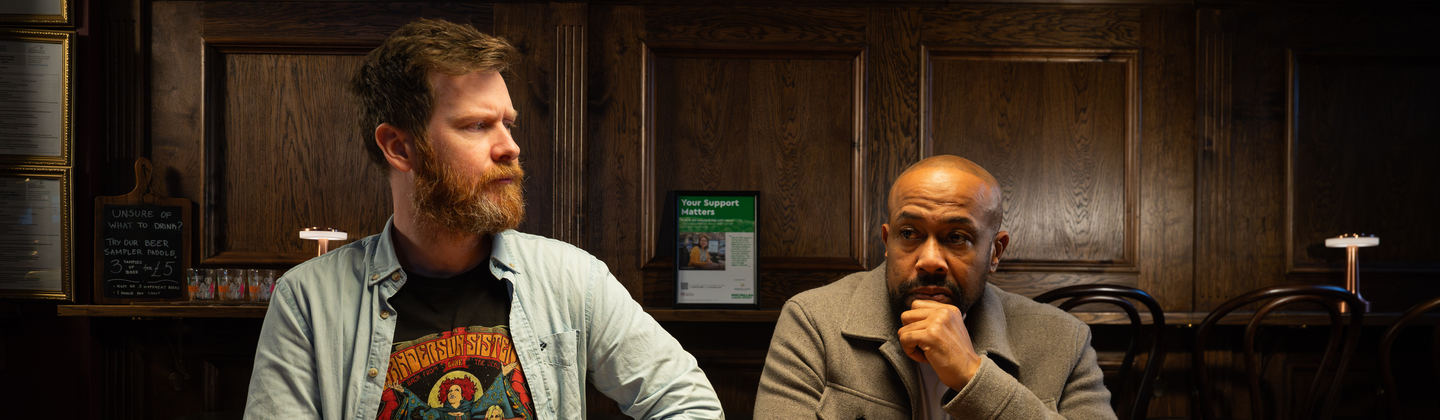 Two men are sitting side by side at a table in a pub. The man on the left is looking over to the man on the right. On the table are drinks and food. 