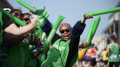 A Macmillan volunteer is cheering at the London Marathon. They have both arms in the air and are holding Macmillan branded balloons. They are wearing a long dark blue top and a Macmillan branded vest on top of it. The volunteer also has on a headscarf and sunglasses. There are other people cheering near them at the event.
