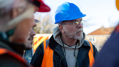 A tradesperson with a blue hard hat and a orange high visibility vest. They are listening to other people talk. They have a short grey beard and are also wearing thin frame glasses. 