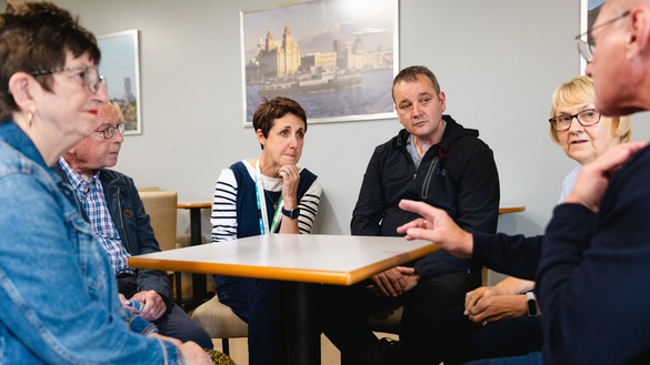 A group of people are sitting down around a square table. People are listening to one person in the group talking. They appear to be indoors.