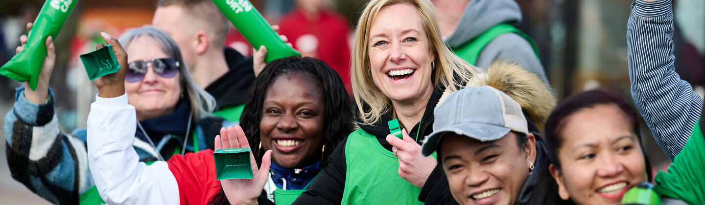 A diverse group of smiling Macmillan Cancer Support volunteers wearing green vests cheer and wave at runners.