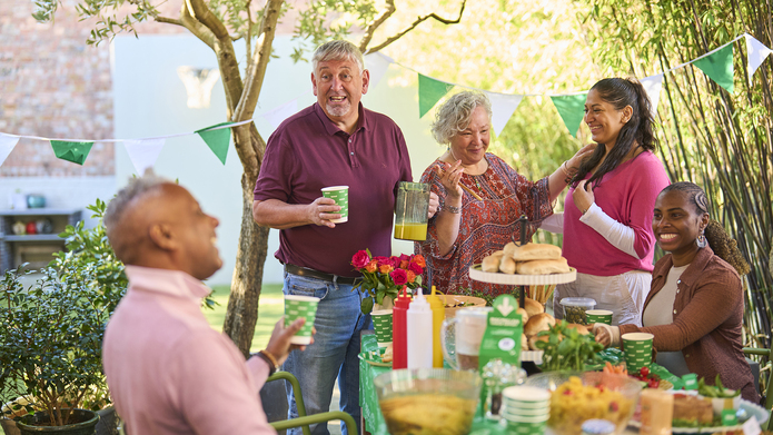 A Macmillan Coffee Morning hosted under the trees in a sunny garden. 5 adults talk and laugh around a table covered in delicious food.