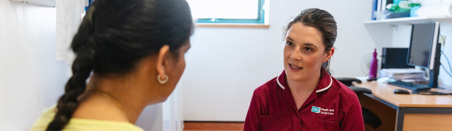 A nurse in a maroon uniform is speaking with a patient and completing a form. The patient has long braided hair and wearing a yellow top.