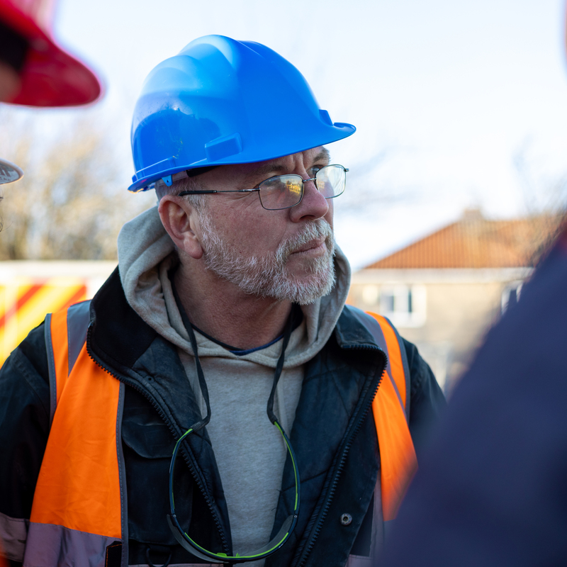 A tradesperson with a blue hard hat and a orange high visibility vest. They are listening to other people talk. They have a short grey beard and are also wearing thin frame glasses. 