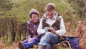 An older couple sitting together on a rocky mound in the country looking at a guidebook.