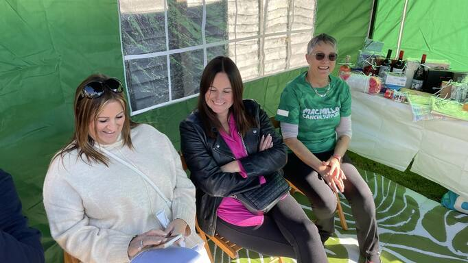 A group of 3 people are sitting together. Two of the people are looking at one persons phone and smiling. Another is wearing a green Macmillan branded top.