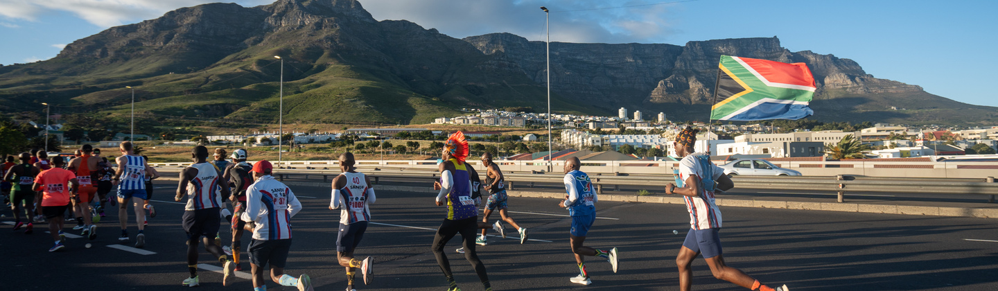 A group of runners are taking part in a marathon. They are running along a road. One person running in the back is holding a South African flag. Beyond the road is a hilly landscape. ©Mark Sampson