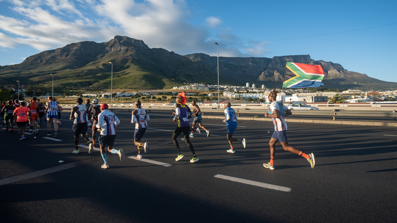 A group of runners are taking part in a marathon. They are running along a road. One person running in the back is holding a South African flag. Beyond the road is a hilly landscape. ©Mark Sampson