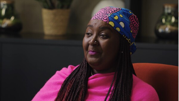 A Black woman sitting in a chair. She has braids and is wearing a brightly coloured headscarf and top.