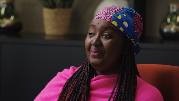 A Black woman sitting in a chair. She has braids and is wearing a brightly coloured headscarf and top.