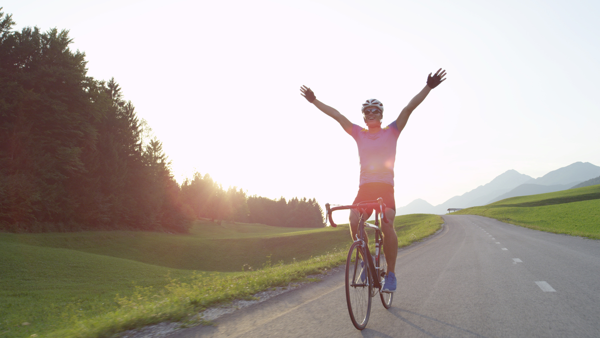 A man cycling alone along an empty country road with his hands up in the air in celebration.