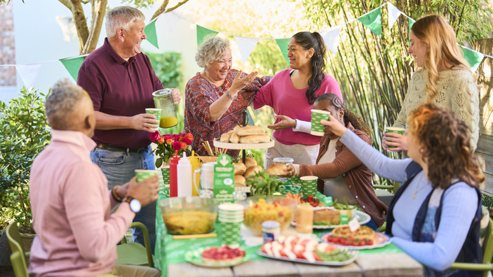 A group of people are gathered together around a picnic table in a garden. The table and garden are decorated for a Coffee Morning. On the table is different food and drink. 