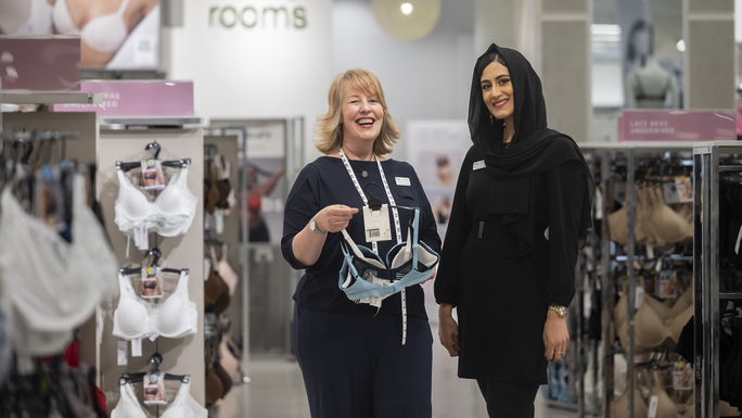 Two colleagues are standing together in the lingerie section in an M&S store. They are standing side to side and the person on the left is holding up a selection of bras. The person on the left has short blond hair. The person on the right is wearing a black head scarf.