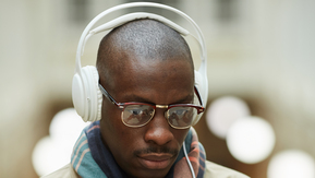 Man is wearing a beige coat and checked scarf, he has white headphones and is looking at his phone,
