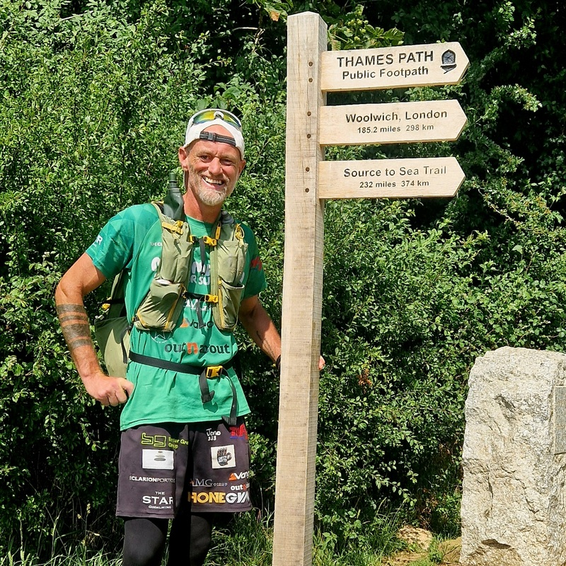 Ben is standing next to a signpost. He is wearing different running gear including a running vest and trainers. The signpost shows the names of different locations and paths. 