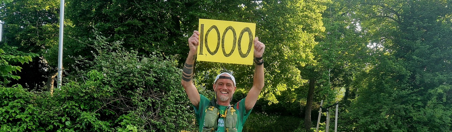 Ben is standing with his arms in the air holding a yellow sign that says 1000 on it. He is outside and is wearing different running gear. 