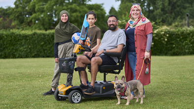 Mohammed with his family outside in a grassy field. Mohammed is wearing a grey top and dark shorts. He is sitting on a mobility scooter.