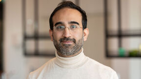 Man is smiling, he is wearing glasses and a cream jumper. He has dark hair and a short beard