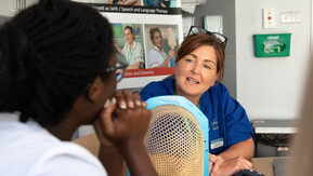 Two people are sitting at a table in a clinic. One is wearing a healthcare professional uniform. There is a radiotherapy mask on the table between them.
