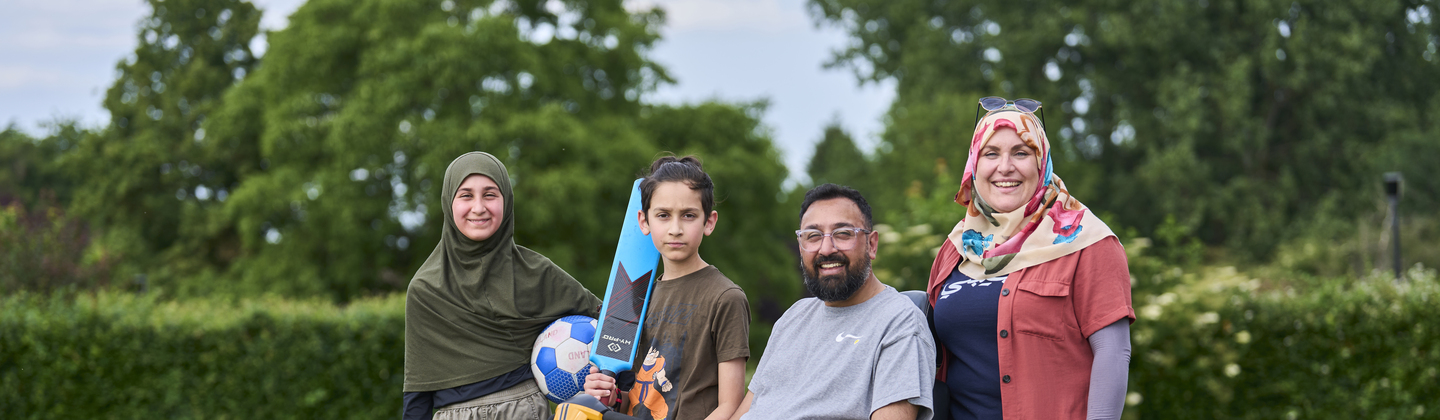 Mohammed with his family outside in a grassy field. Mohammed is wearing a grey top and dark shorts. He is sitting on a mobility scooter.
