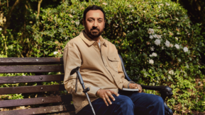 A man is sitting on a park bench. He has a walking stick next to him and a book in his hand.