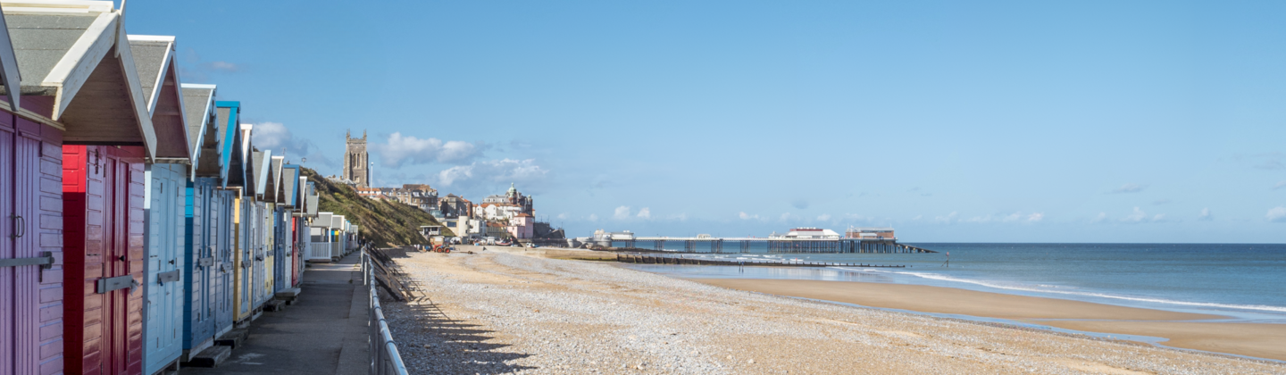 A beach in Norfolk on a sunny day with colourful beach huts and a pier.