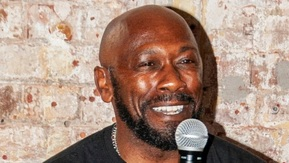 Charlie is sitting on a chair holding a microphone and smiling. He has a short beard and is wearing a black tshirt.