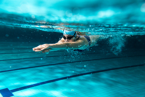 A woman swims with her hands out in front having just dived into the pool. She wears a swim cap and goggles.