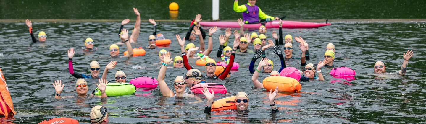 A group of swimmers are in a lake. They are waving. Many have floating devices attached to their body. There is also a person in a kayak in the background waving.
