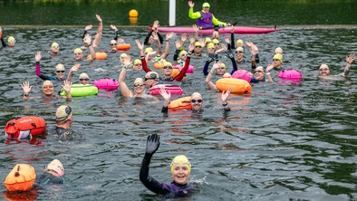A group of swimmers are in a lake. They are waving. Many have floating devices attached to their body. There is also a person in a kayak in the background waving.