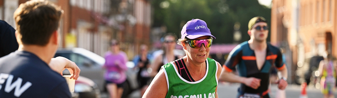 A runner in a Macmillan branded top taking part in an event. They are also wearing a purple hat and black and pink shorts. They are running on a road closed off for the event.