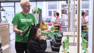 A hairdresser is styling a client's hair. The hairdresser is wearing a green Macmillan branded top and is using a round hair brush and blow dryer to style the client's hair. The client is sitting down and is holding a white coffee cup in their hands. The salon is decorated for a Coffee Morning.