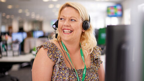 Professional working on the Macmillan Support Line, she is wearing headphones with a mic attached. She is has blonde hair and is wearing a floral print top with a green lanyard.