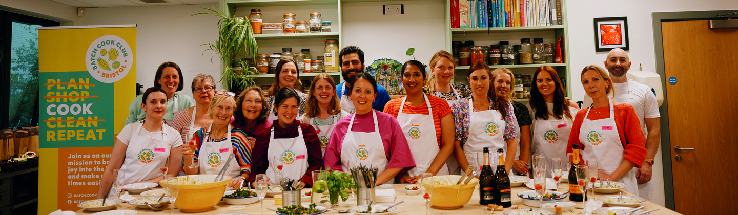 Members of the Batch Cook Club are standing together for a group photo. In front of them is a table with used plates and glasses. The members are wearing white cooking aprons. They are inside a cooking class kitchen. Photography: Rebecca Naen