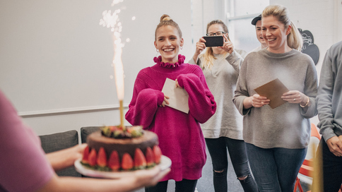 People are standing in an office and watching someone walk over holding a chocolate cake. The decadent cake is covered in strawberries. Many people are holding cards in their hands. One person is taking a photo with their phone. Photograph: SolStock/Getty Images
