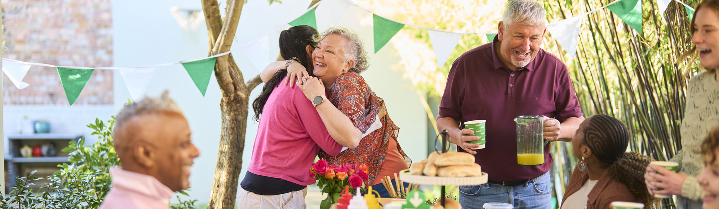 A group of people are outside and having a Coffee Morning. They are gathered around a table that is full of tasty bites and drinks. Two people are hugging at one end of the table.