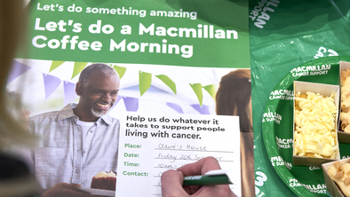 A person writing on a Coffee Morning poster with the details of their event. The poster is on a table decorated for a Coffee Morning. 