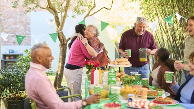 A group of people are outside and having a Coffee Morning. They are gathered around a table that is full of tasty bites and drinks. Two people are hugging at one end of the table.