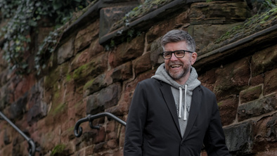 Phil is walking down a stair case alongside a brick wall. He has short hair and is wearing black frammed glasses, a dark jacket, grey hoodie and dark trousers. 