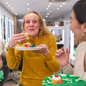 A person in a bright yellow top has taken a bite out of a colourful sweet treat. They are holding up a plate with other sweet bites on it and fruit. Two people next to them have turned to watch them enjoy their food.