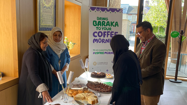 Two people are standing behind a table that has many different sweet treats and other food on sale for a Coffee Morning. The people are representatives from the Baraka Khan Foundation. On the other side of the table two people are looking at the different food on sale. There is a sign near them that says 'Bring Baraka To Your Coffee Morning.'