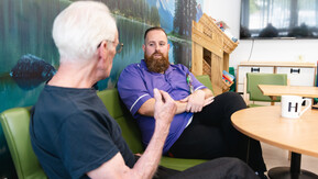 An person with white hair sits across from a healthcare professional in a purple uniform. There is a round table in front of them