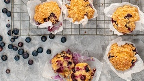Golden-brown blueberry muffins with a crumb topping rest on a cooling rack, wrapped in white parchment paper. Scattered blueberries and visible moist interiors make these treats look irresistibly fresh and delicious.