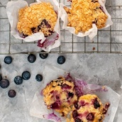 Golden-brown blueberry muffins with a crumb topping rest on a cooling rack, wrapped in white parchment paper. Scattered blueberries and visible moist interiors make these treats look irresistibly fresh and delicious.