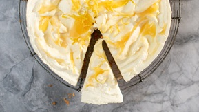A creamy lemon cheesecake with swirls of glossy lemon curd and delicate strips of lemon zest sits on a wire rack. One slice is cut and slightly pulled out, revealing its rich, velvety interior.