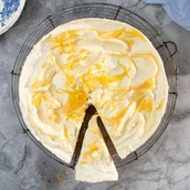 A creamy lemon cheesecake with swirls of glossy lemon curd and delicate strips of lemon zest sits on a wire rack. One slice is cut and slightly pulled out, revealing its rich, velvety interior.