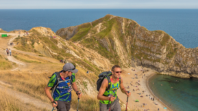 Two hikers walking a path along the cliff with Durdle Door in the background